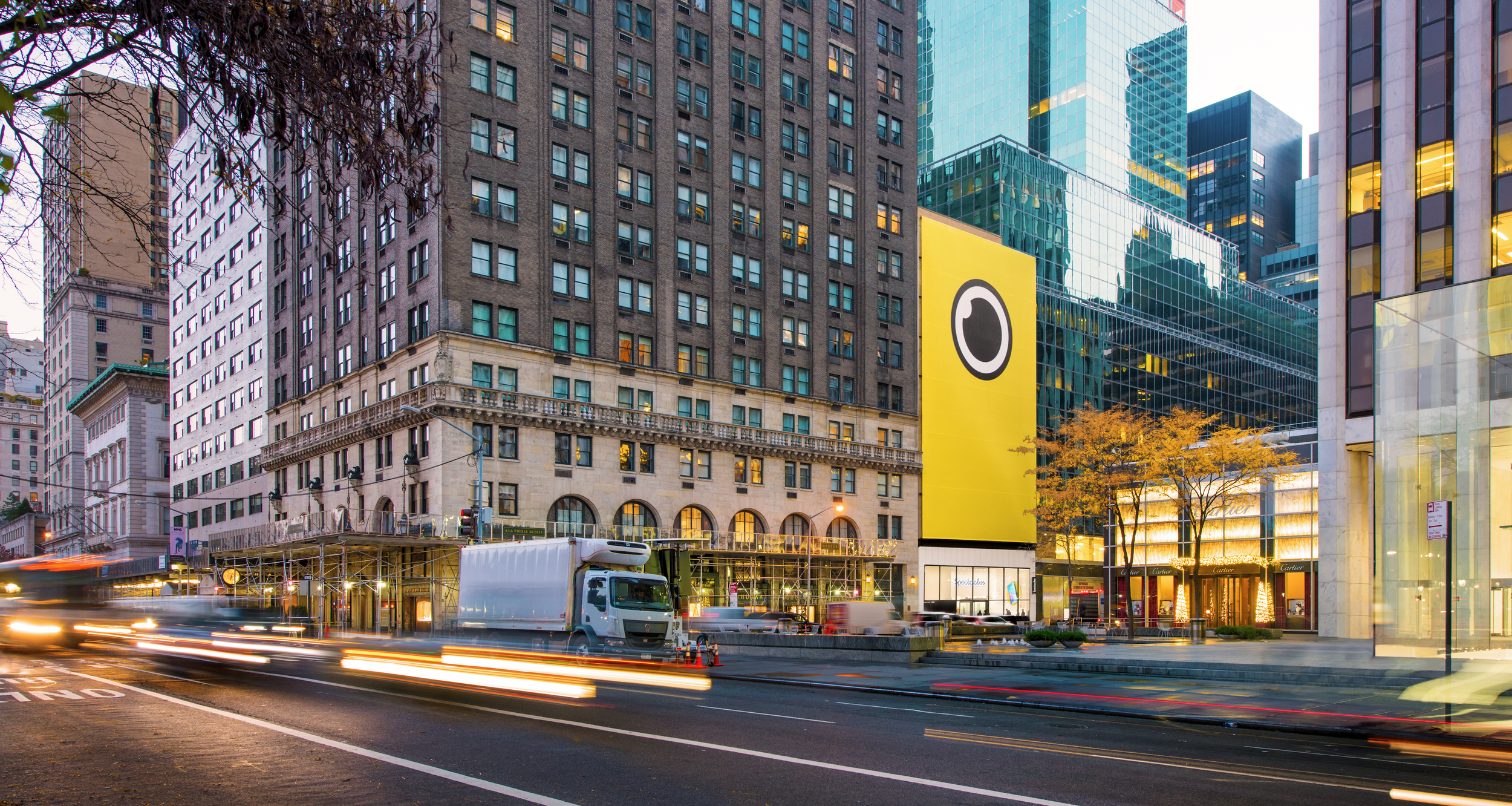 Dusk Manhattan corner with a giant yellow Spectacles camera-lens facade above a ground-floor store, Cartier next door